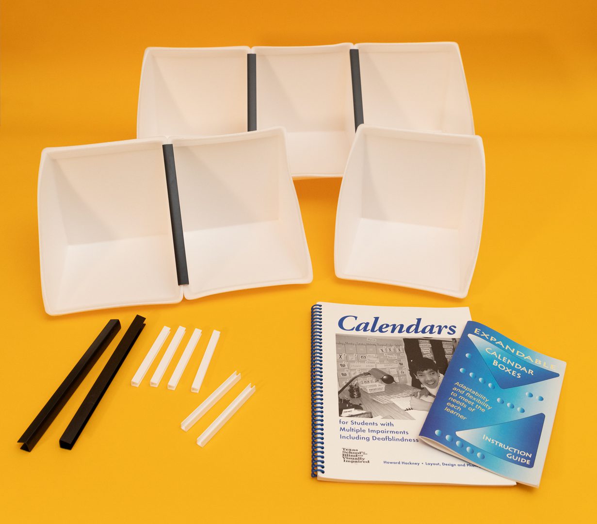 White_Expandable-Calendar-Boxes-Kit_above-shot_web All components of the White Expandable Calendar Boxes kit laid out on a yellow background. Included are six white foam boxes attached to one another with black and white U-channels, two additional large black U-channels, six smaller white U-channels, and the print guidebooks placed in front.
