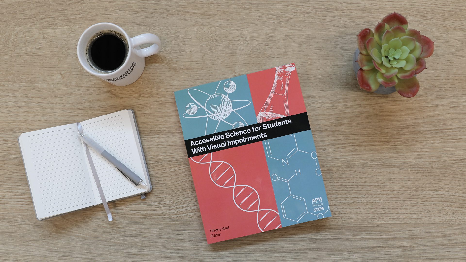 Accessible Science On Table An overhead shot of a copy of Accessible Science sitting on a wooden surface next to a mug of black coffee, a notebook and pen, and a green and red succulent.