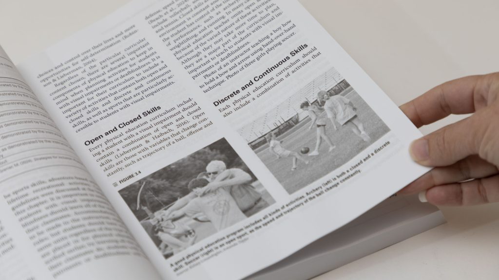 A close up of a text book page shows images of students played soccer, and a boy shooting a bow and arrow. The surrounding sections of text are on building physical skill sets.