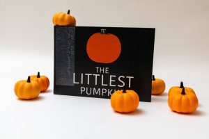 The Littlest Pumpkin storybook stood up on its long edge showing its cover with the title and a simple illustration of an orange pumpkin. Small decorative pumpkins are positioned around and on top of the book.