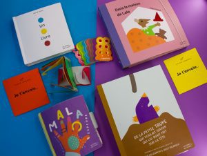 Multiple tactile books lay against a blue and purple backdrop. Some of them are small and hard, while some are bigger and made of felt material.