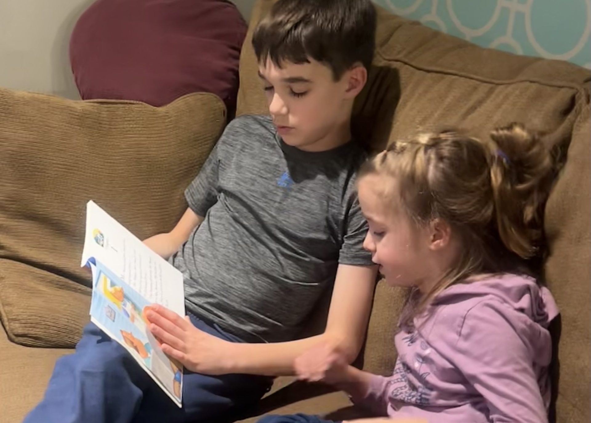 Two students, a boy and a girl sit ont he couch together reading the braille book.